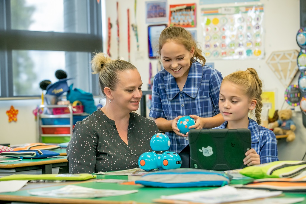 Teacher sitting at a desk with two primary school aged children. One student is showing the teacher the iPad. The other is standing behind them, holding an educational resource ball.